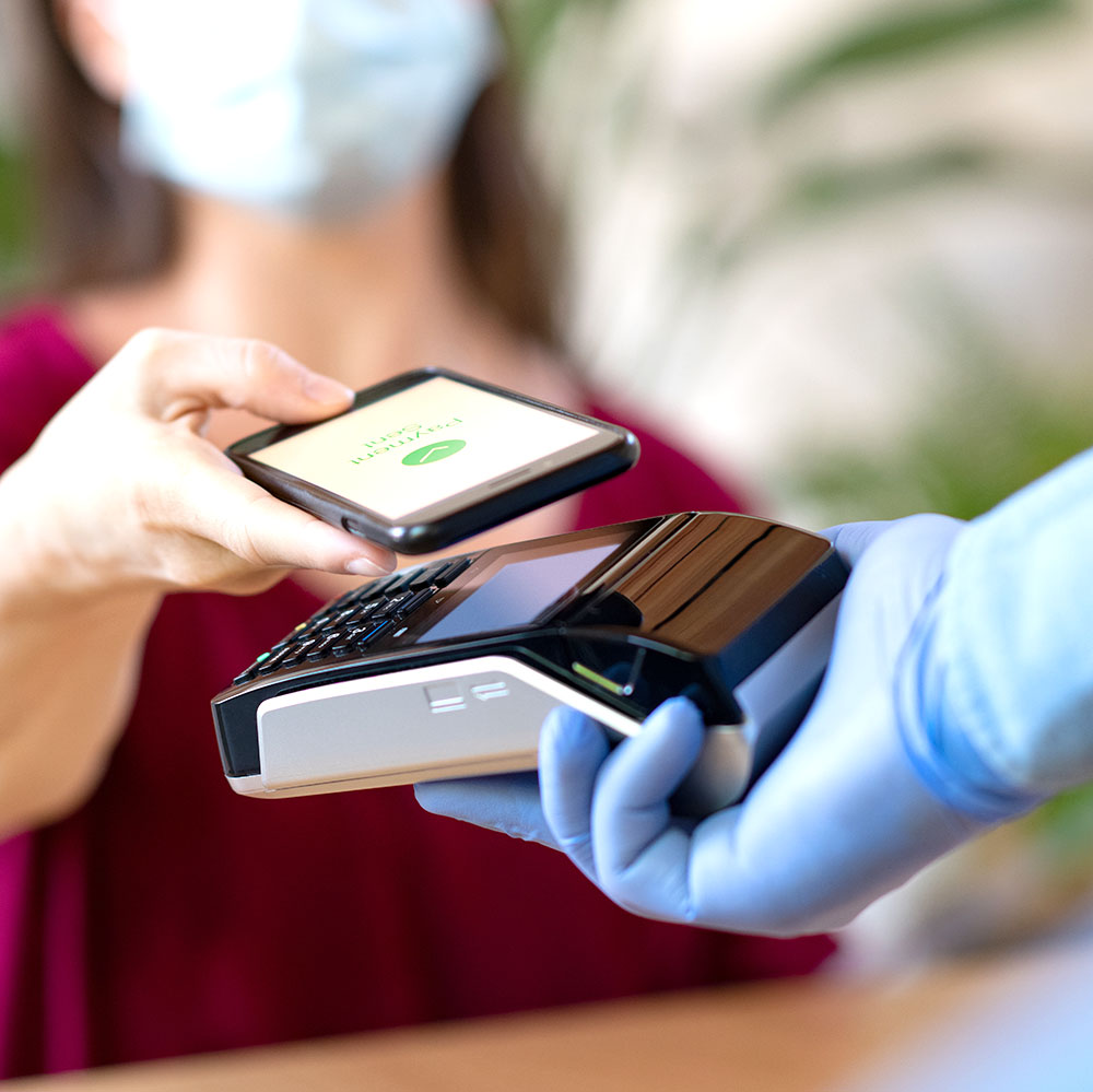 Cashier hand holding credit card reader machine and wearing disposable gloves while client holding phone for NFC payment.